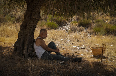 Adult man in white tank top and jeans taking rest under olive tree in summer