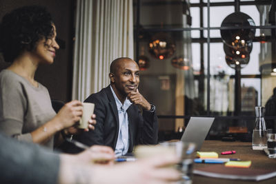 Smiling businessman sitting with hand on chin by colleagues in meeting room