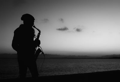 Silhouette man playing musical instrument while standing by lake against sky