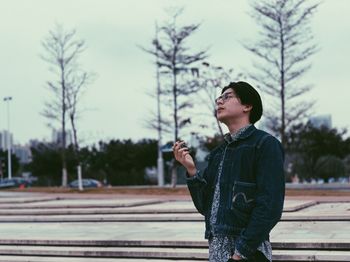 Young man smoking cigarette while standing against sky