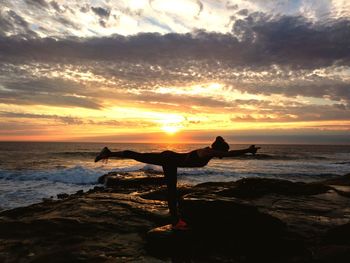 Side view of woman practicing yoga on rocks by sea against sky during sunset