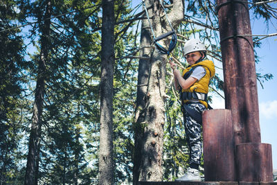 Happy boy attached on a hanging rope getting ready for zip lining on rope course in adventure park.