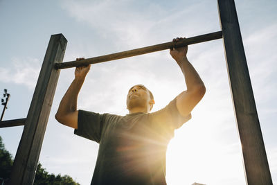Active sportsman exercising on monkey bar against sky during sunny day