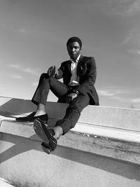Low angle portrait of young man wearing suit sitting on retaining wall against sky