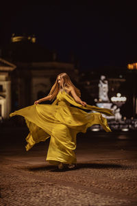Woman wearing yellow evening gown while spinning on footpath in city at night