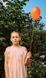 A girl in a pink dress and a red ball in her hand stands on the street on a summer day. 