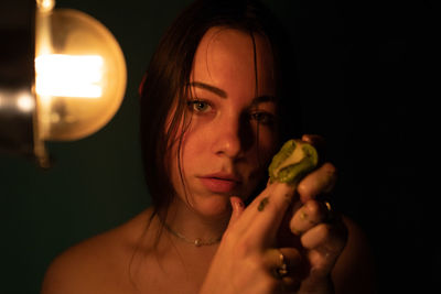 Close-up portrait of young woman holding kiwi by illuminated light bulb