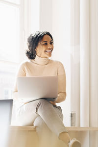 Smiling woman looking away while using laptop at desk in office