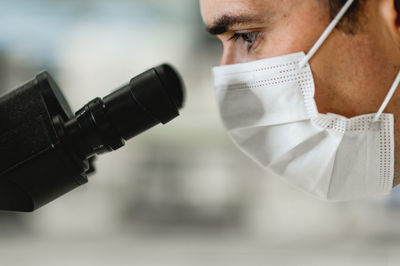 Side view of skilled male scientist in protective mask using microscope while conducting chemical experiment in modern lab