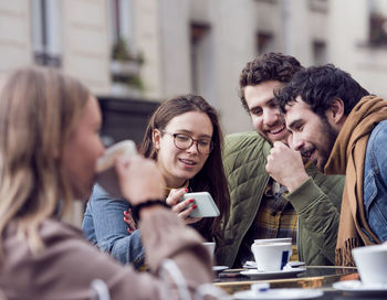 Happy woman showing smart phone to male friends at sidewalk cafe