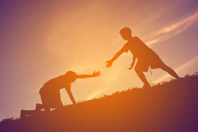 Low angle view of teenage boy helping girl climbing on hill against sky during sunset
