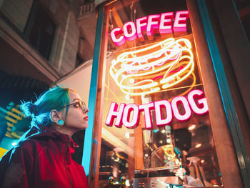 Young woman looking at illuminated sign on store while standing outdoors at night