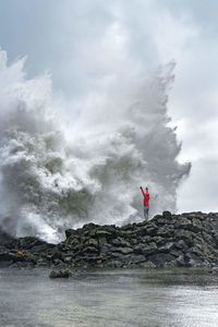 Man standing on rock by splashing sea wave against sky