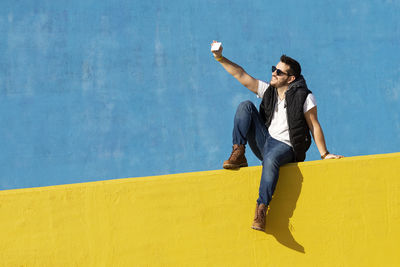 Young bearded man with sunglasses sitting on yellow wall,taking selfie