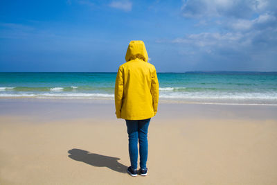 Rear view of woman wearing yellow hooded jacket at beach against sky on sunny day