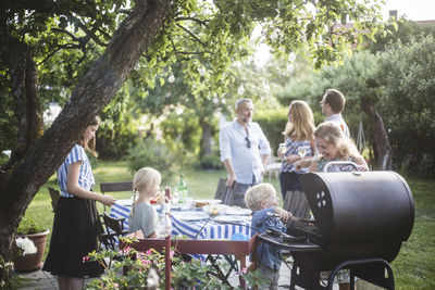 Boy preparing food on barbecue grill with mother while family enjoying at dining table in backyard party