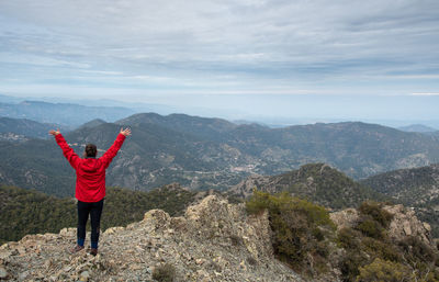 Woman with raised arms standing on rocky top against a cloudy sky enjoying mountain range panorama.