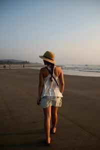 Full length rear view of woman walking at beach against clear sky during sunset