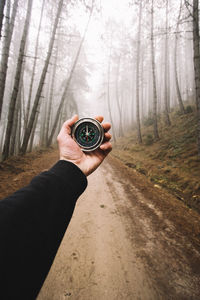 Close-up of hand holding navigational compass on road against bare trees in forest during foggy weather