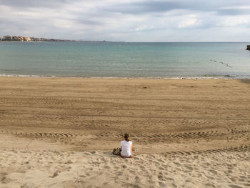 Rear view of woman sitting at beach against sky