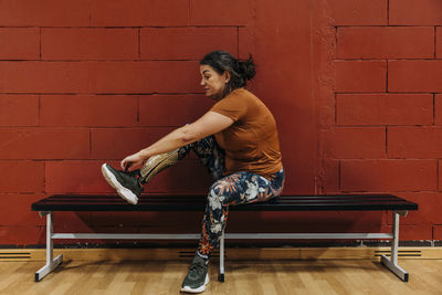 Side view of athlete with disability wearing shoe while sitting on bench near red wall at sports court