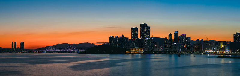 Illuminated modern buildings by bay against sky during sunset