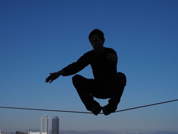 Low angle view of silhouette man performing stunt on rope against clear blue sky