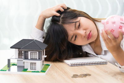 Irritated woman holding piggy bank with model home on table