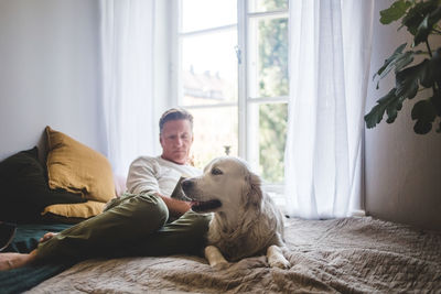 Dog resting on bed while senior man reading book against window at home