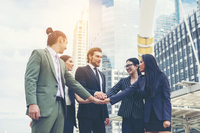 Happy young colleagues stacking hands against buildings in city 