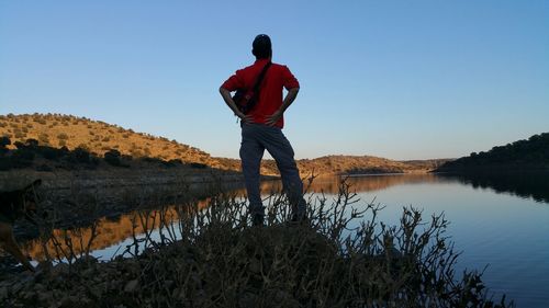 Rear view of man standing by lake against clear sky during sunset