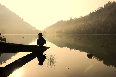 Silhouette man crouching by lake against sky during sunset