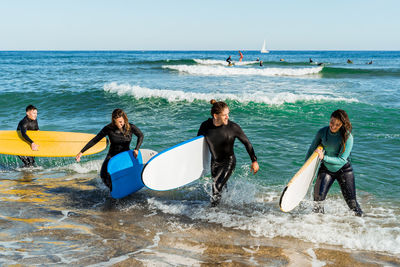 Group of young friends in swimsuits with surfboards walking out wavy ocean after surfing together in summer day
