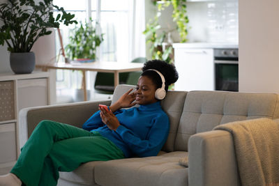 Happy african american woman in headphones listening to music using mobile phone lies on sofa