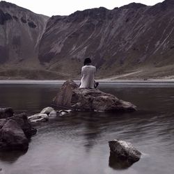 Rear view of woman sitting on rock amidst lake against mountain