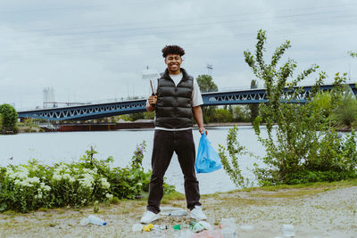 Portrait of smiling young man collecting garbage near river against sky