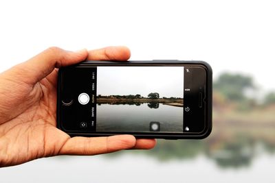 Cropped hand photographing lake against clear sky