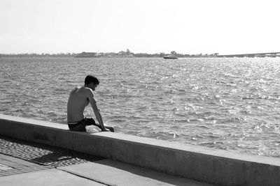 Shirtless man sitting on retaining wall at pier