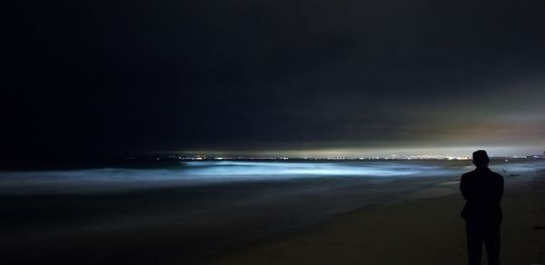 Rear view of man standing at beach against sky at night