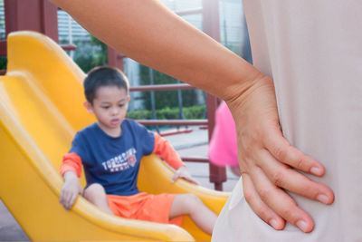 Cropped image of mother with hand on hip by son playing on slide