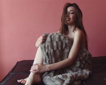 Portrait of beautiful woman covered in blanket sitting on bed against wall