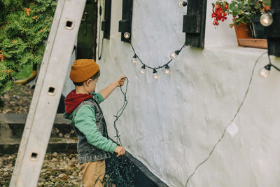 Boy holding string lights standing outside house