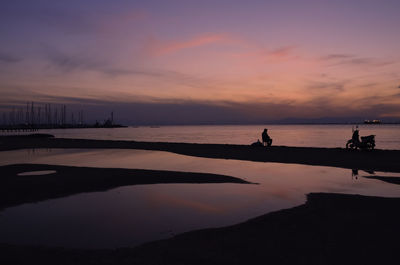 Scene in beach with silhouettes of sails, a chapel,a man sitting and a motorcycle at purple sunset