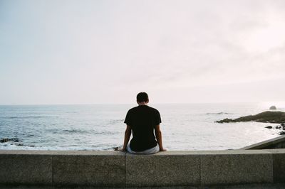 Rear view of man sitting on retaining wall while looking at sea against sky