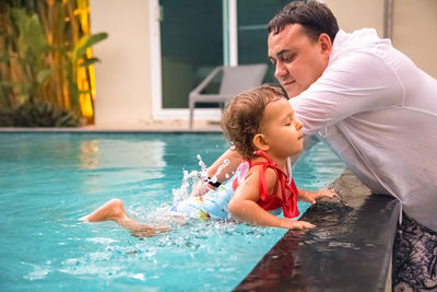 Father teaches cute toddler little girl in swimsuit to swim in the pool holding his hand.