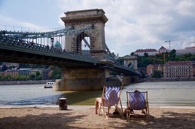 People sitting on deck chairs at banks of danube against chain bridge