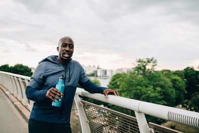 Portrait of thirsty sportsman holding water bottle while standing by railing on footbridge against sky