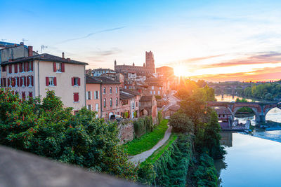 Trees and buildings by river against sky during sunset