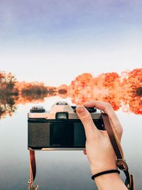 Cropped hand of photographer photographing calm lake against sky during autumn