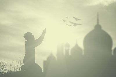 Silhouette boy praying against mosque during sunset